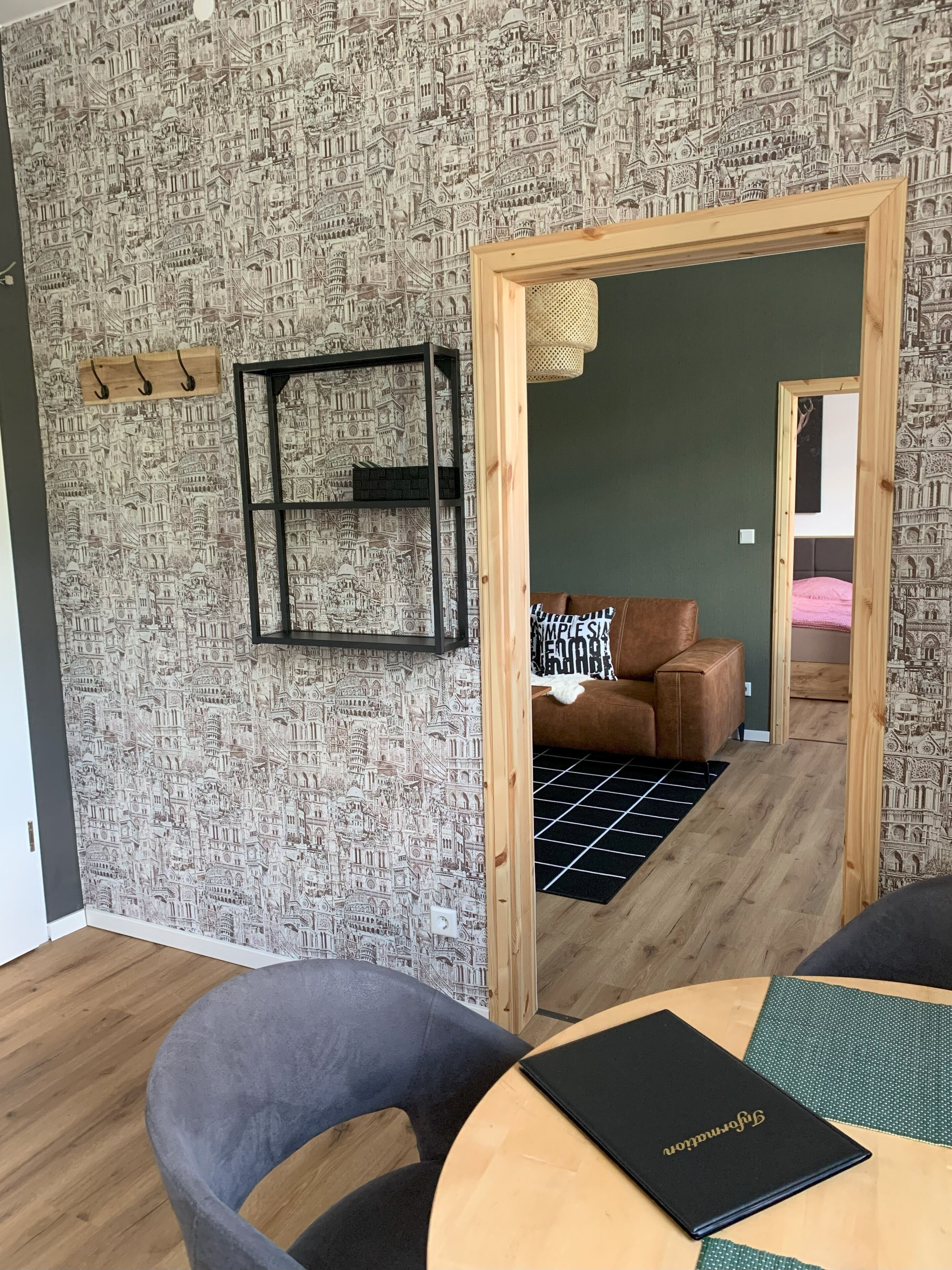 A modern room with a patterned wallpaper, a wooden door leading to another room, a round table with a black folder, and a grey chair.