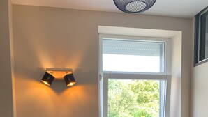 A cozy dining area with a round table, two chairs, a window with blinds, a wall-mounted shelf with kitchen utensils, and a framed picture.