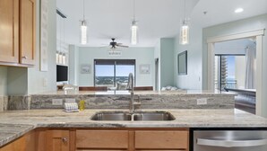 A modern kitchen with a granite countertop, stainless steel sink, wooden cabinets, and a ceiling fan. - Origin 1231 - One Bedroom - Sunrise and Gulf Views (Panama City Beach)