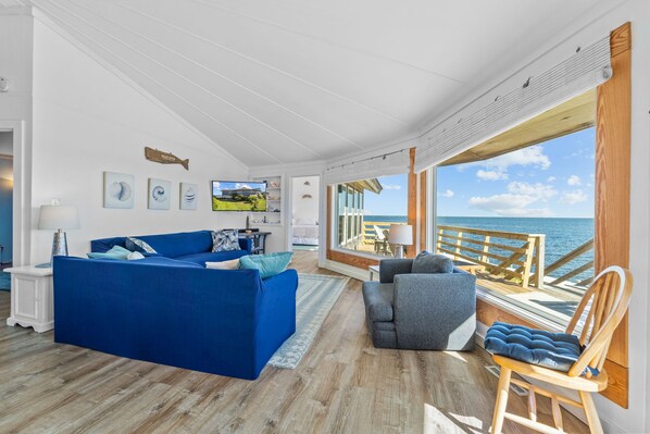 A spacious living room with a blue sofa, a grey armchair, and a wooden deck overlooking the sea.