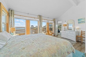 A bedroom with a large bed, a dresser, a chair, and a view of the water through large windows.
