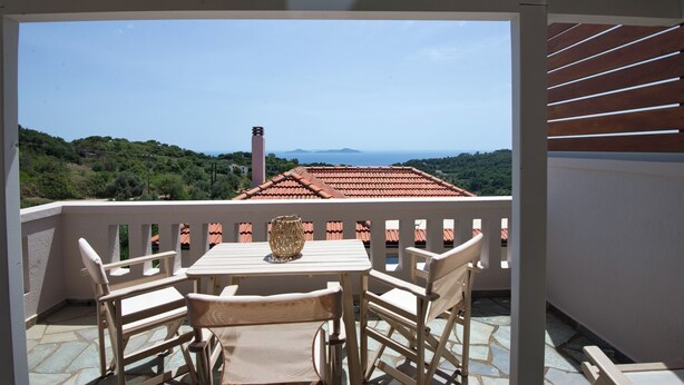A balcony with a table and chairs, overlooking a mountainous landscape and a red-roofed building. - Likavgies Comfort Maisonettes (Alonissos)