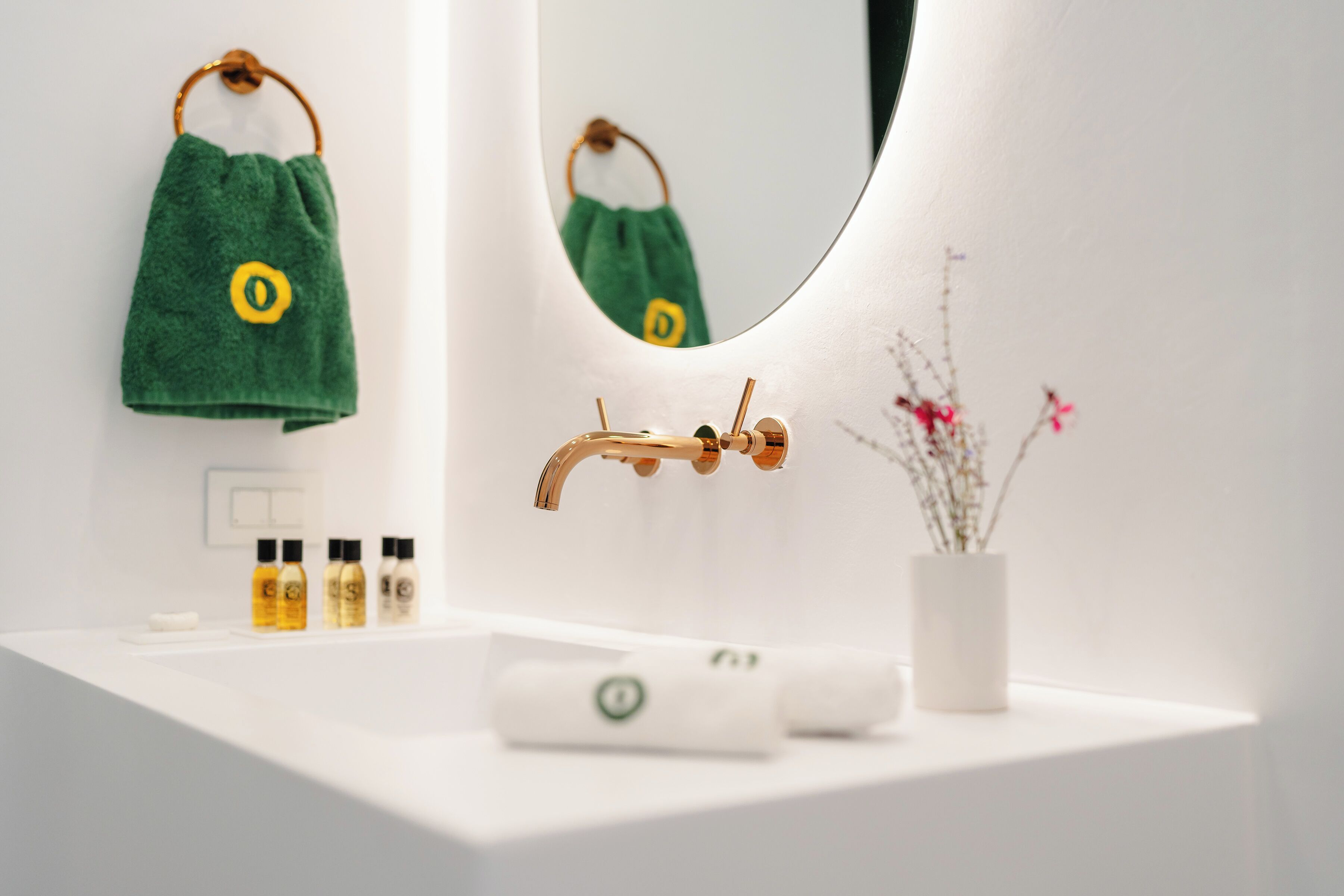 Un baño con un lavabo blanco, una toalla verde colgada en la pared, un espejo y un jarrón con flores.
