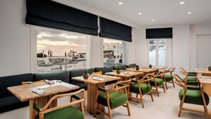 A modern dining area with wooden tables and green upholstered chairs, large windows with black blinds, and a view of the sea and buildings. - Croco Mykonos (Mykonos)