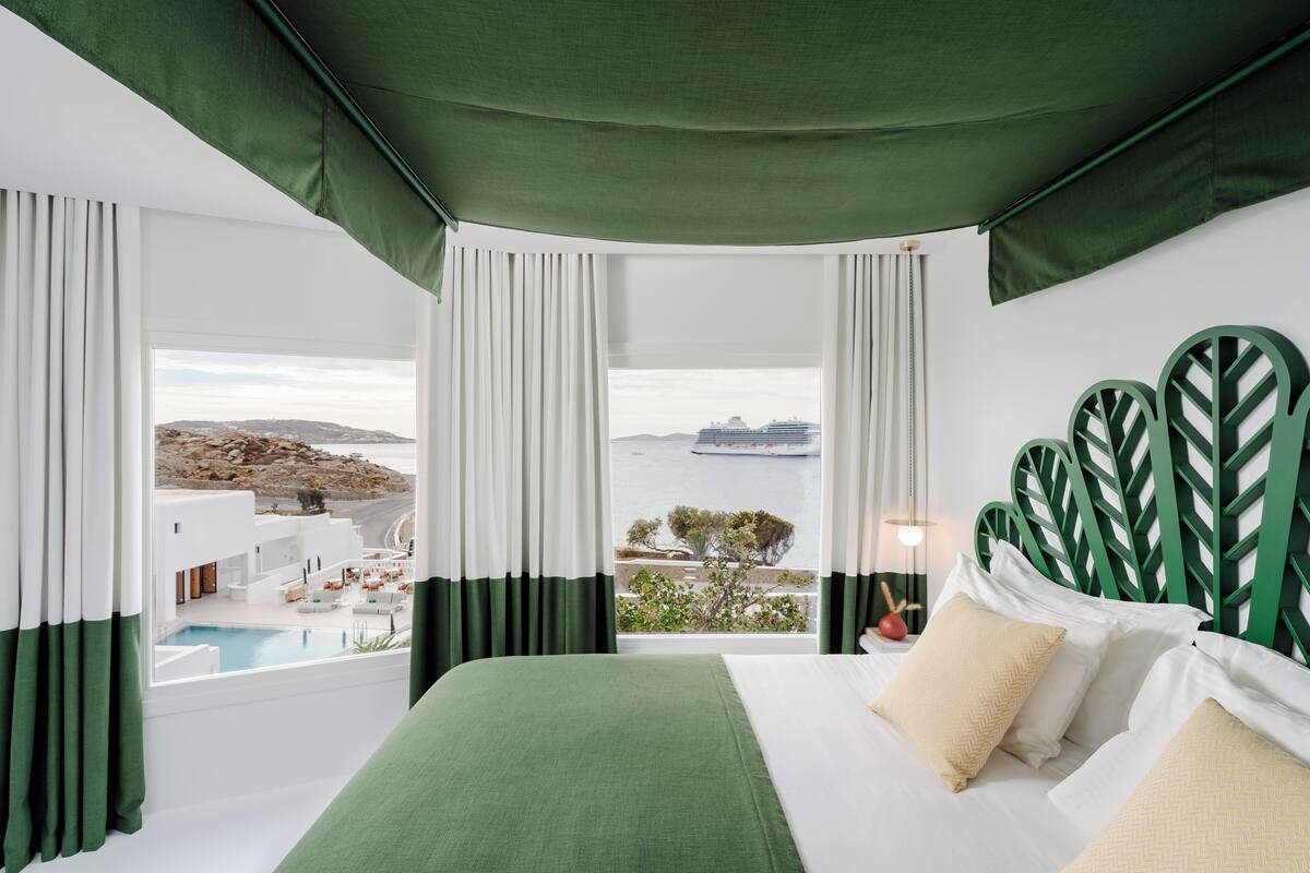 A hotel room with a bed, a view of a pool, and a balcony overlooking a body of water and buildings.