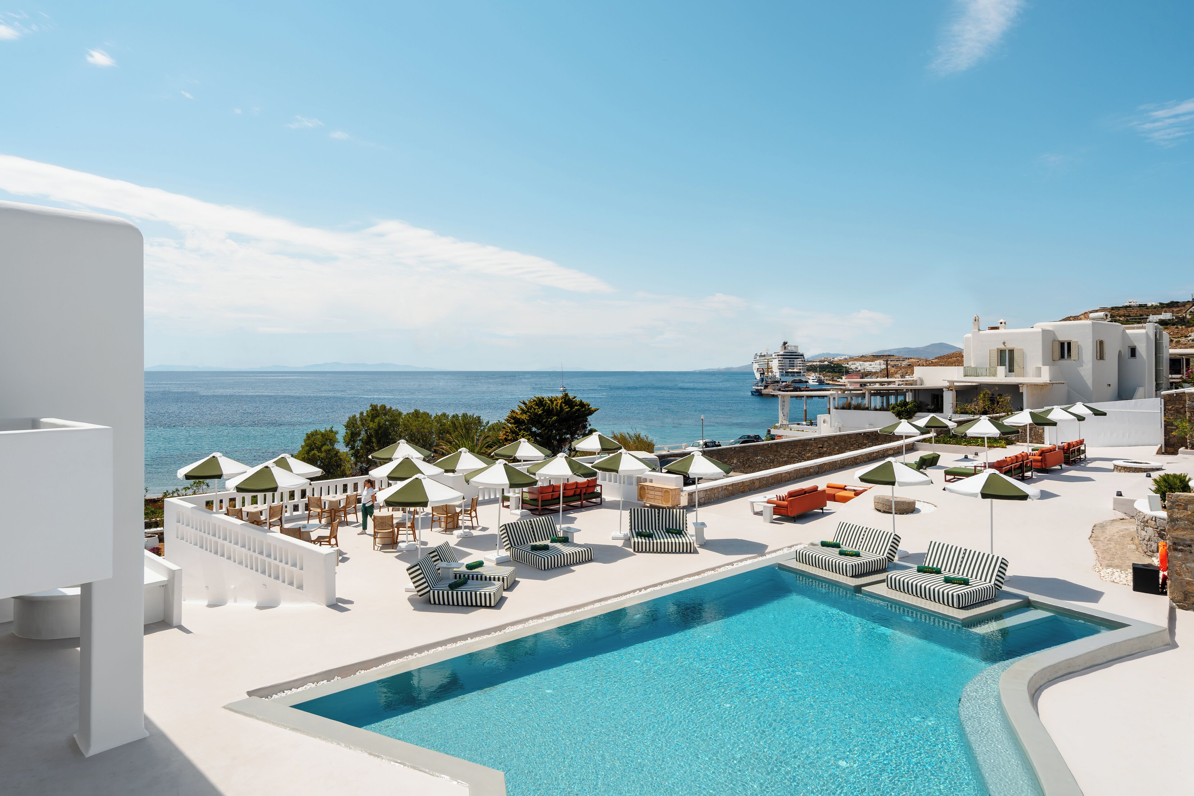 A poolside area with lounge chairs and umbrellas, a clear view of the sea, and a building with a white facade.