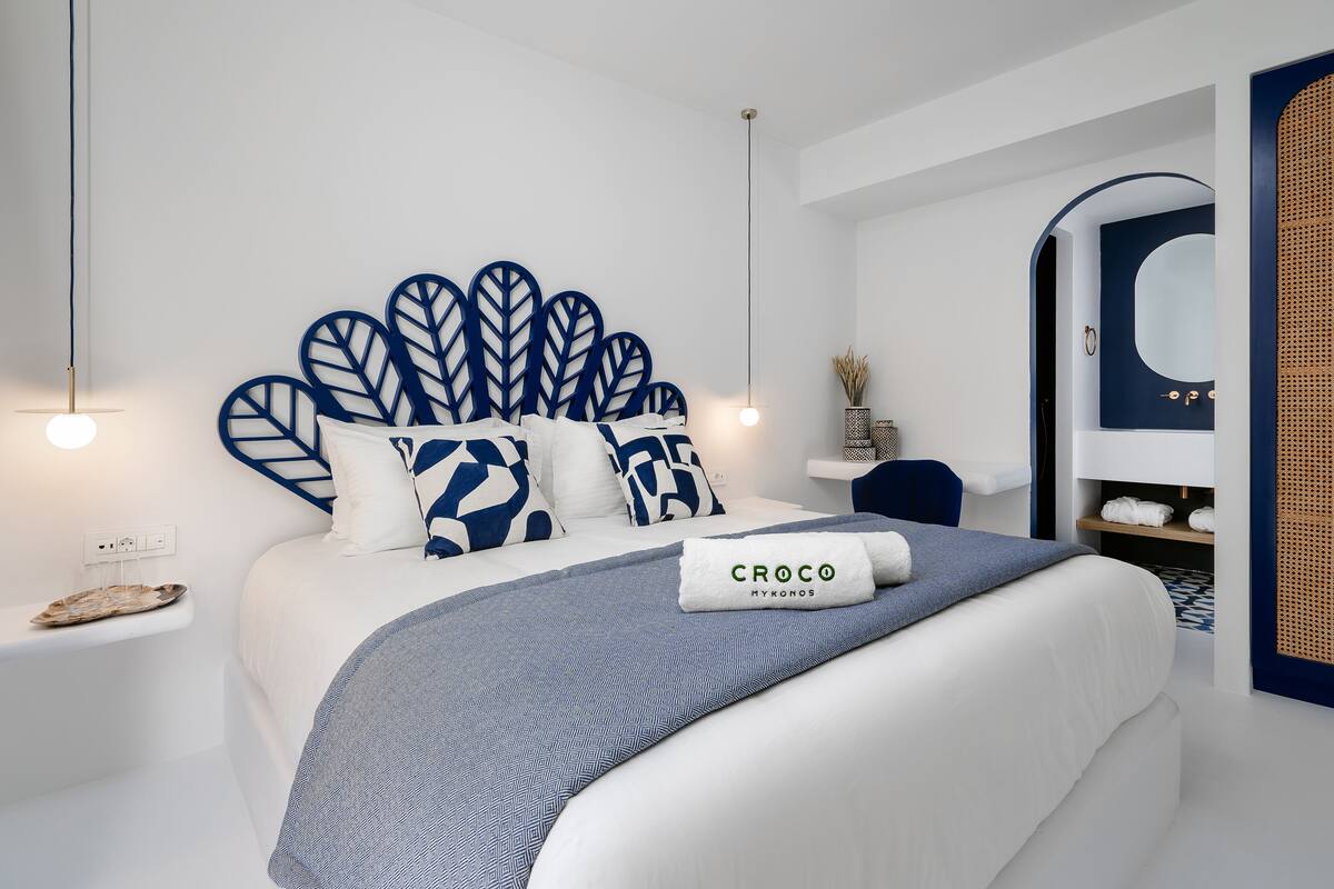 A modern hotel room with a large bed, a headboard with leaf patterns, a bedside table with a lamp, and a bathroom with a mirror and towel rack.