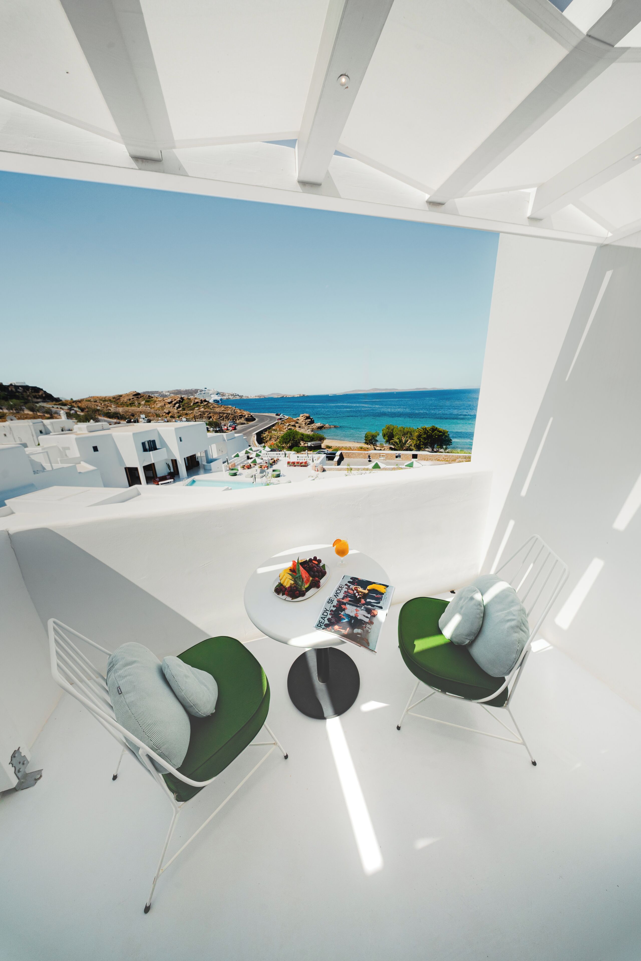 A modern room with a view of the sea, featuring two green cushioned chairs, a small round table with a magazine, and a white ceiling.