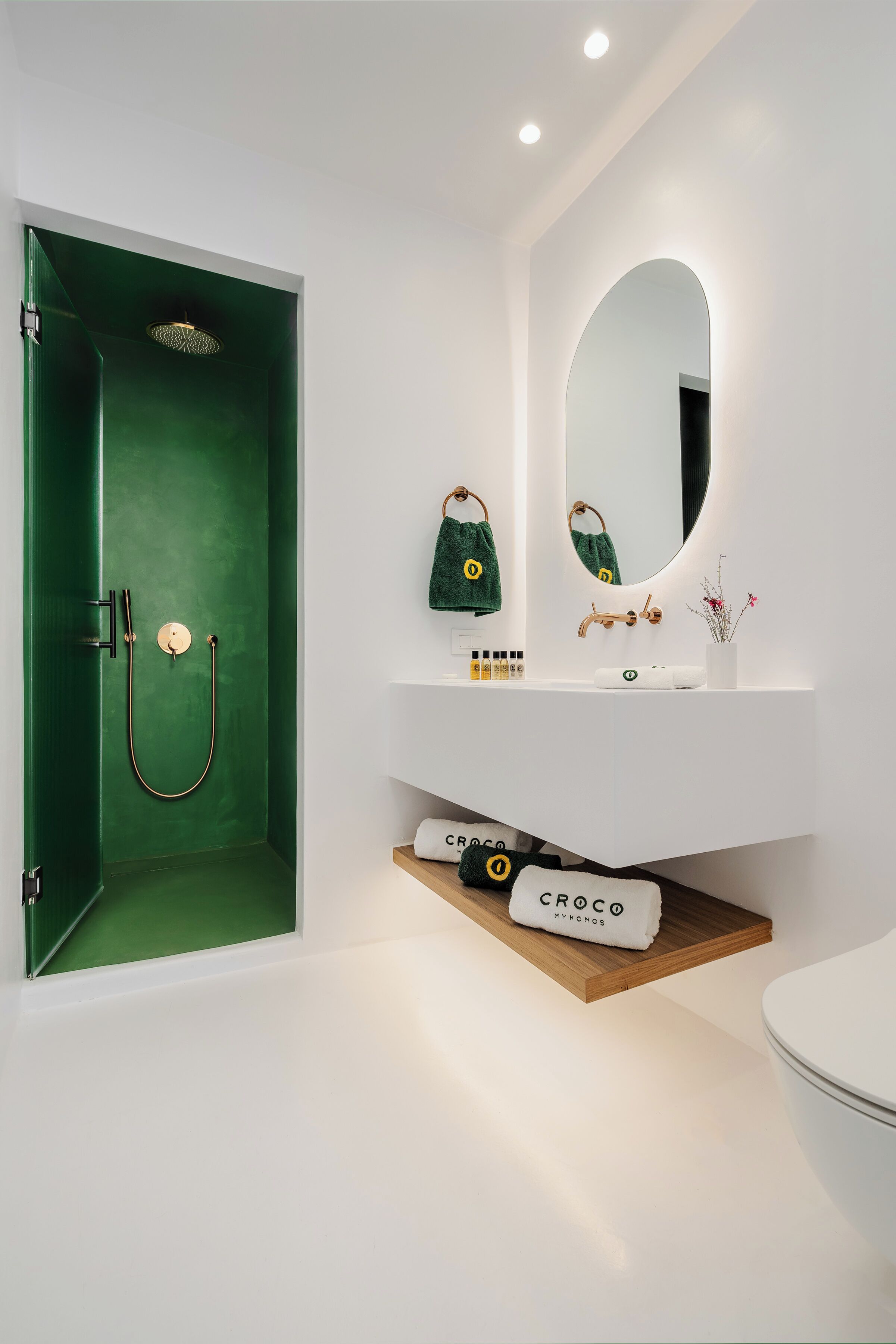 A bathroom with a green shower enclosure, a white sink with a mirror, and a Croco towel on a wooden shelf.