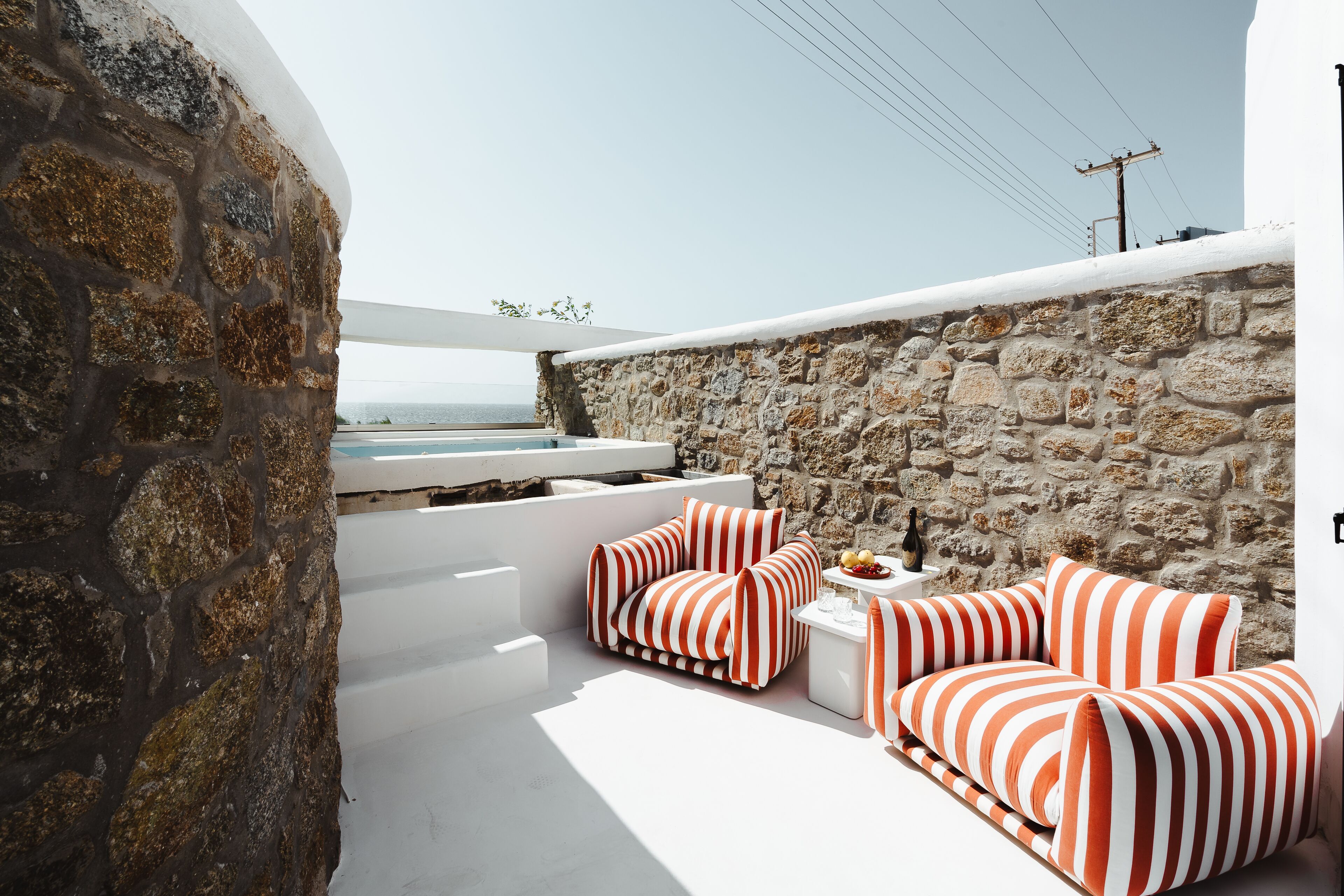A stone wall with a white bench and striped cushions, a small table with a bottle and fruit, and a view of the sea.