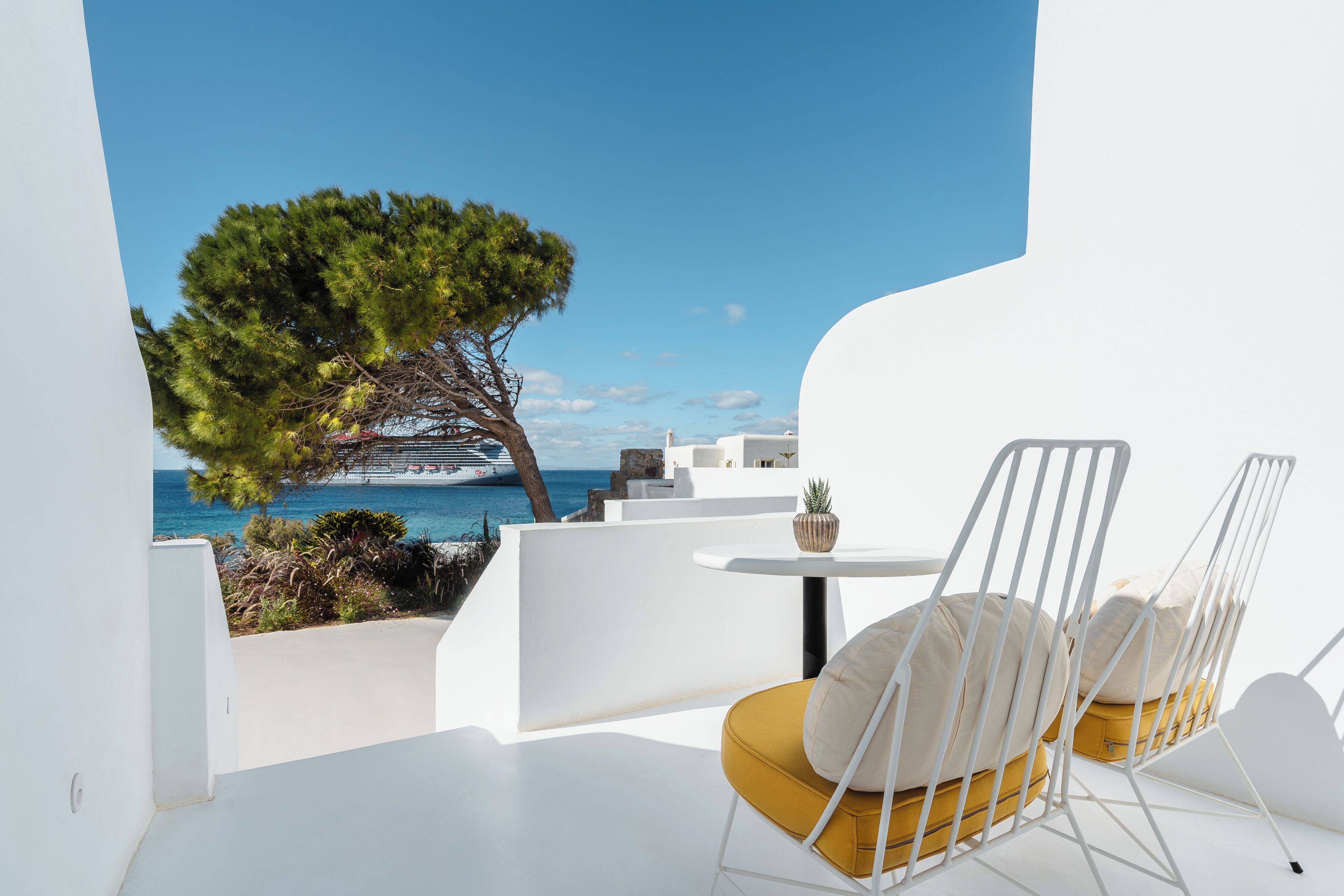 A balcony with white walls, a yellow chair, and a view of the sea and a cruise ship.