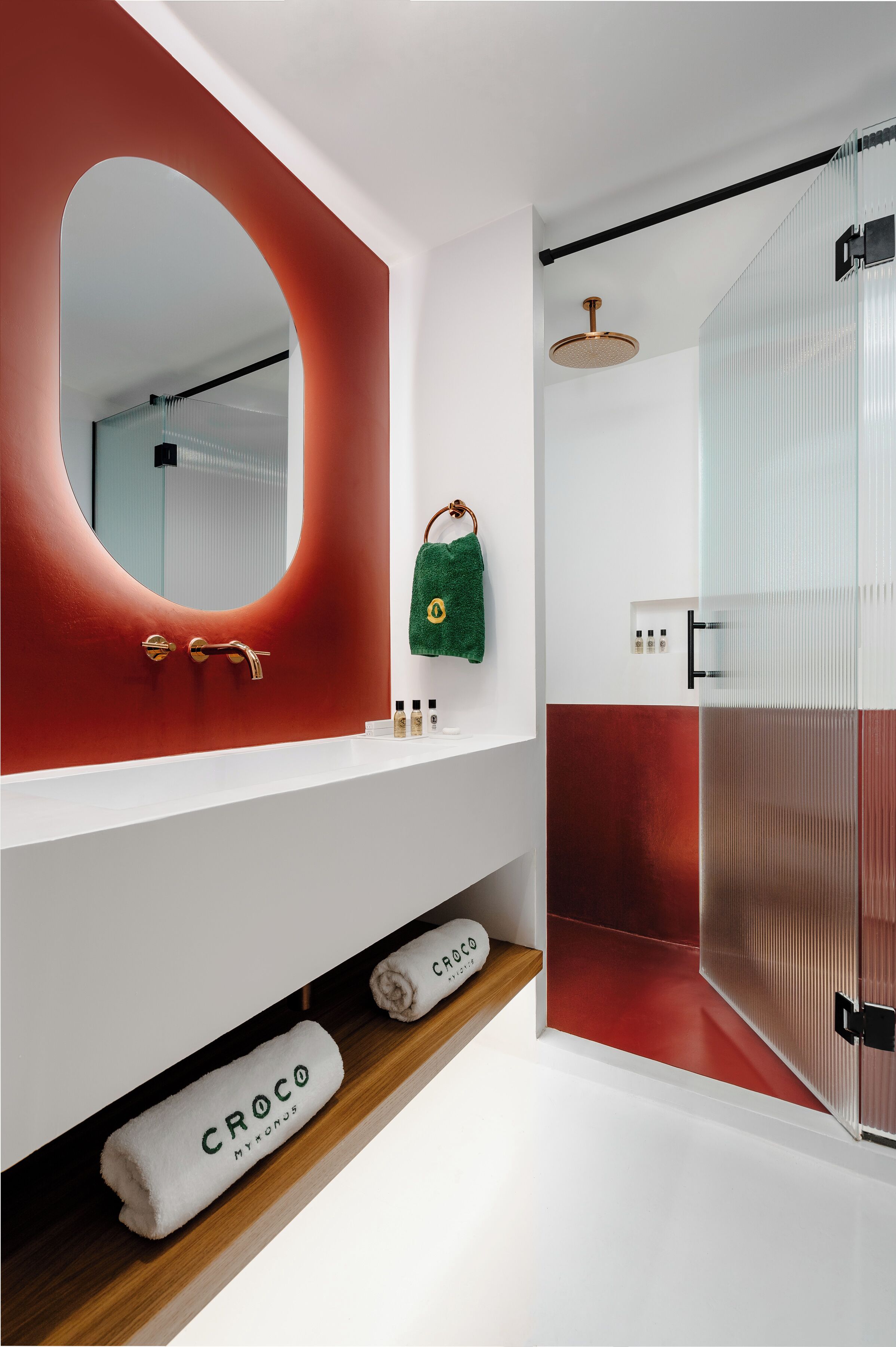 Un baño moderno con una pared de acento roja, un espejo redondo, una toalla verde y un box de ducha.