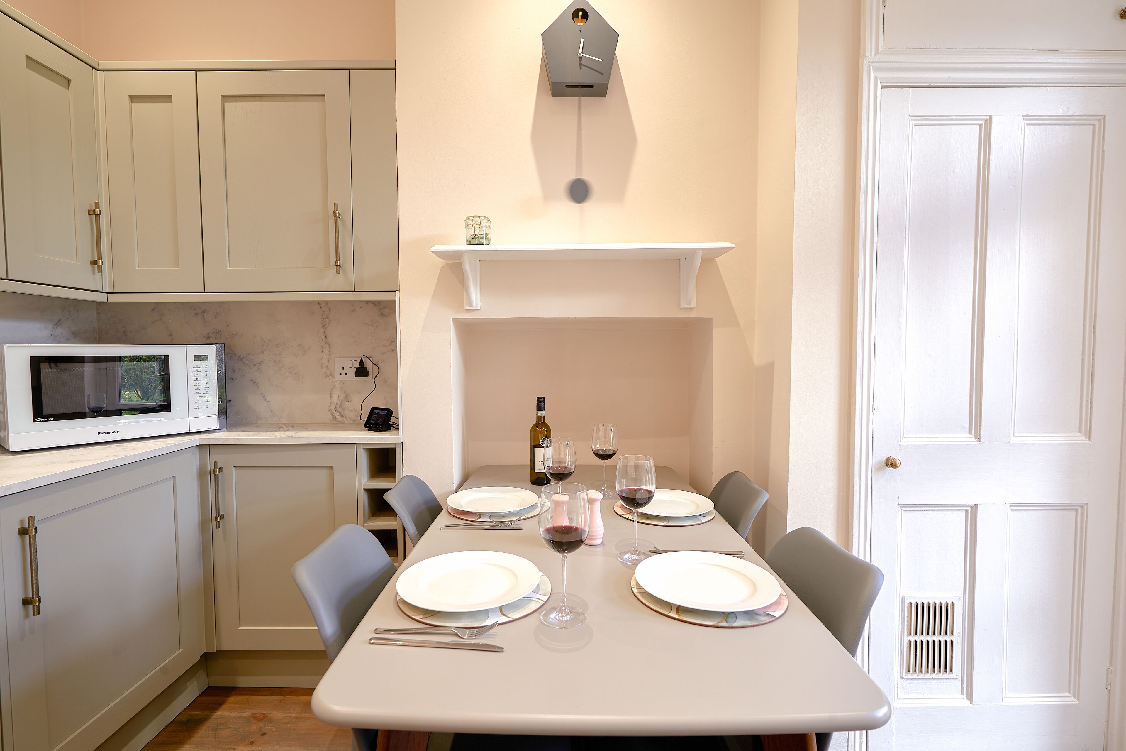 A dining area with a table set for four, a microwave, and a clock on the wall.