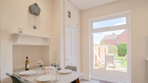 A dining area with a table set for four, a wine bottle, and glasses. There is a white door leading to an outdoor patio with a brick wall and greenery. - A Stunning Ground Floor Apartment (Reydon)