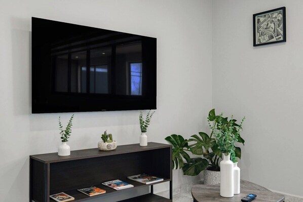 A modern living room with a large flat-screen TV mounted on the wall, a dark wooden console table with decorative plants and magazines, and a round coffee table with a remote control.
