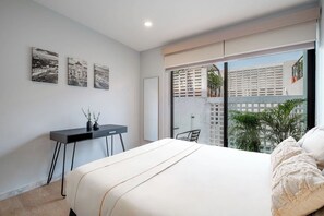 A modern bedroom with a large bed, a desk, and a view of buildings through a sliding door.