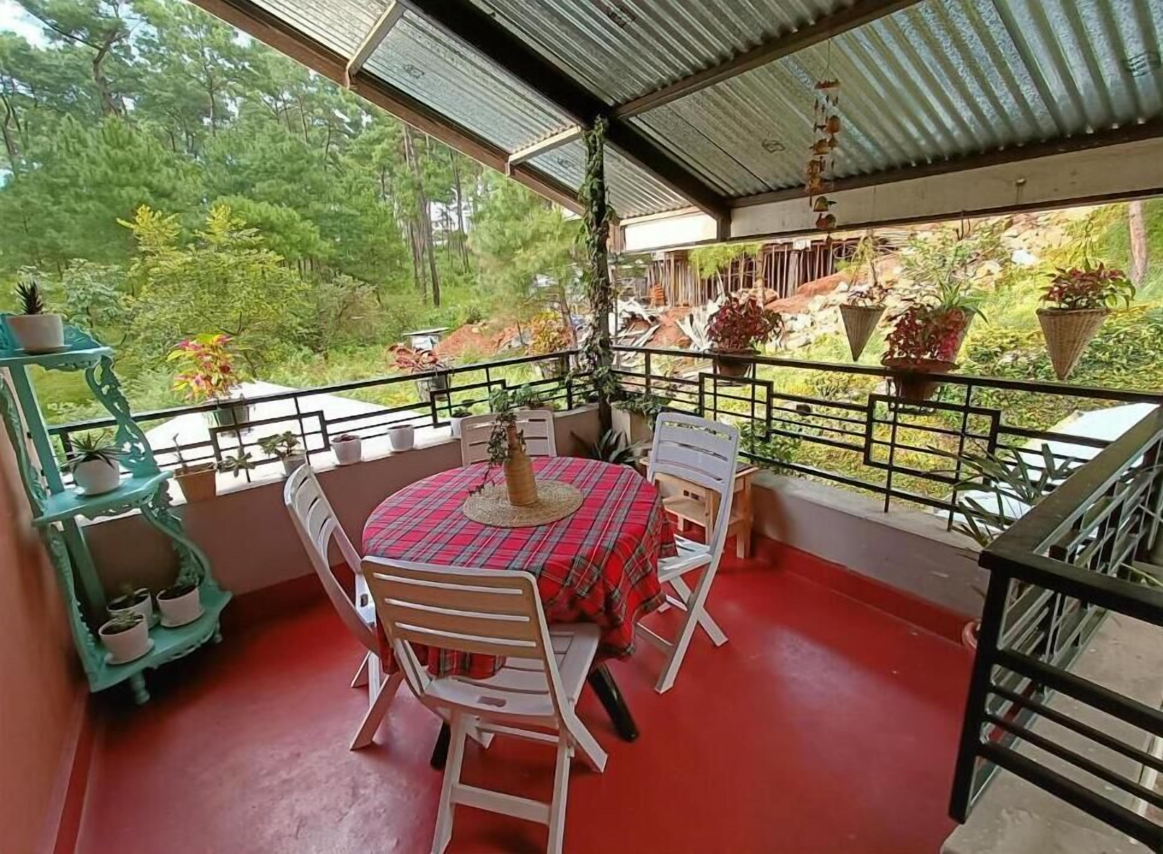 A covered outdoor seating area with a round table and chairs, surrounded by potted plants and a view of greenery.