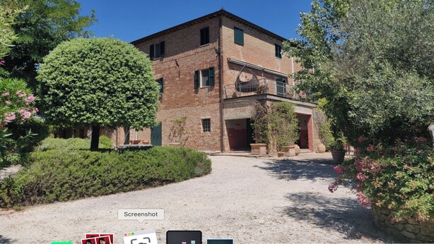 A two-story brick building with a balcony, surrounded by greenery and a gravel path.