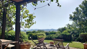 A wooden deck with chairs and a table, surrounded by potted plants and a view of a green landscape with mountains in the distance.