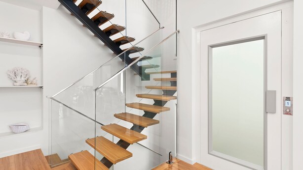 Un escalier en colimaçon moderne, doté de rampes en verre et de marches en bois, mène à une porte blanche avec un panneau de verre.
