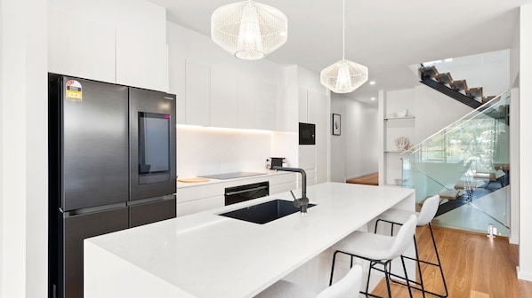 A modern kitchen with a stainless steel refrigerator, a white island with bar stools, and a glass staircase leading to a balcony.