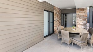 Un patio techado con una mesa redonda y sillas de mimbre, junto a un edificio con revestimiento color beige y detalles de piedra.