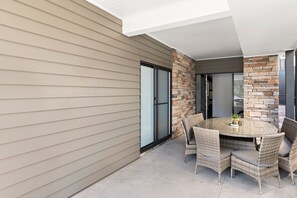 Een overdekt terras met een ronde tafel en rieten stoelen, grenzend aan een gebouw met beige bekleding en stenen accenten.