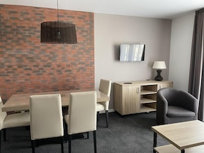 Quarto de hotel com parede de tijolos decorativa, televisão de tela plana, escrivaninha com luminária e área de estar com cadeiras e mesa.