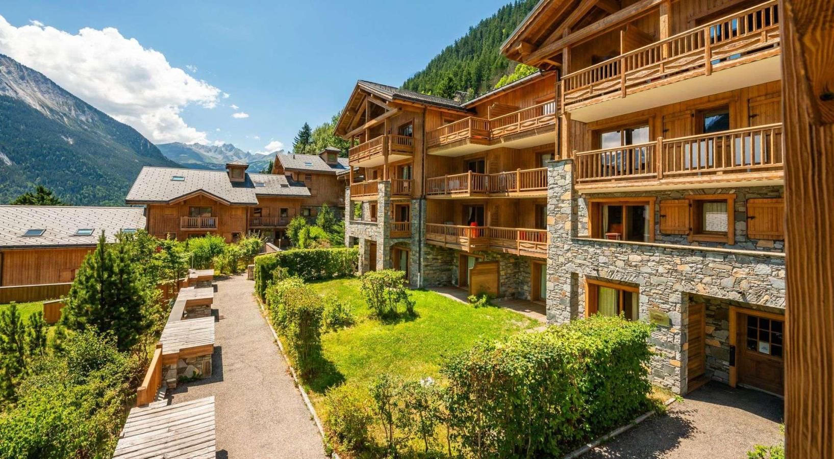 A multi-story wooden building with balconies, surrounded by a well-maintained garden and a clear view of mountains in the background.