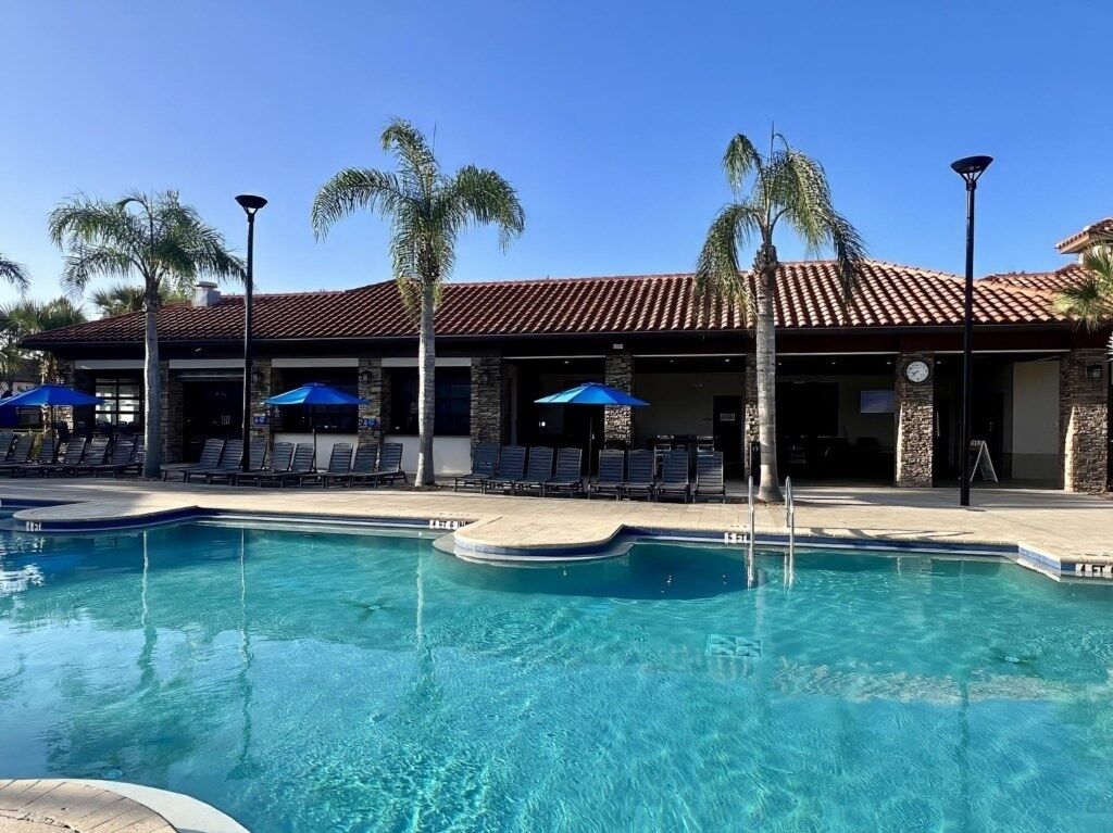 A swimming pool with lounge chairs and umbrellas, a building with a tiled roof, and palm trees.