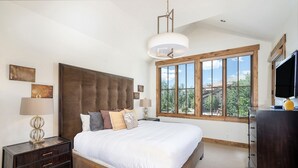 Ein Schlafzimmer mit einem großen Bett, einem Nachttisch, einer Kommode, einem Fernseher und einem Fenster mit Blick auf Bäume und Gebäude.