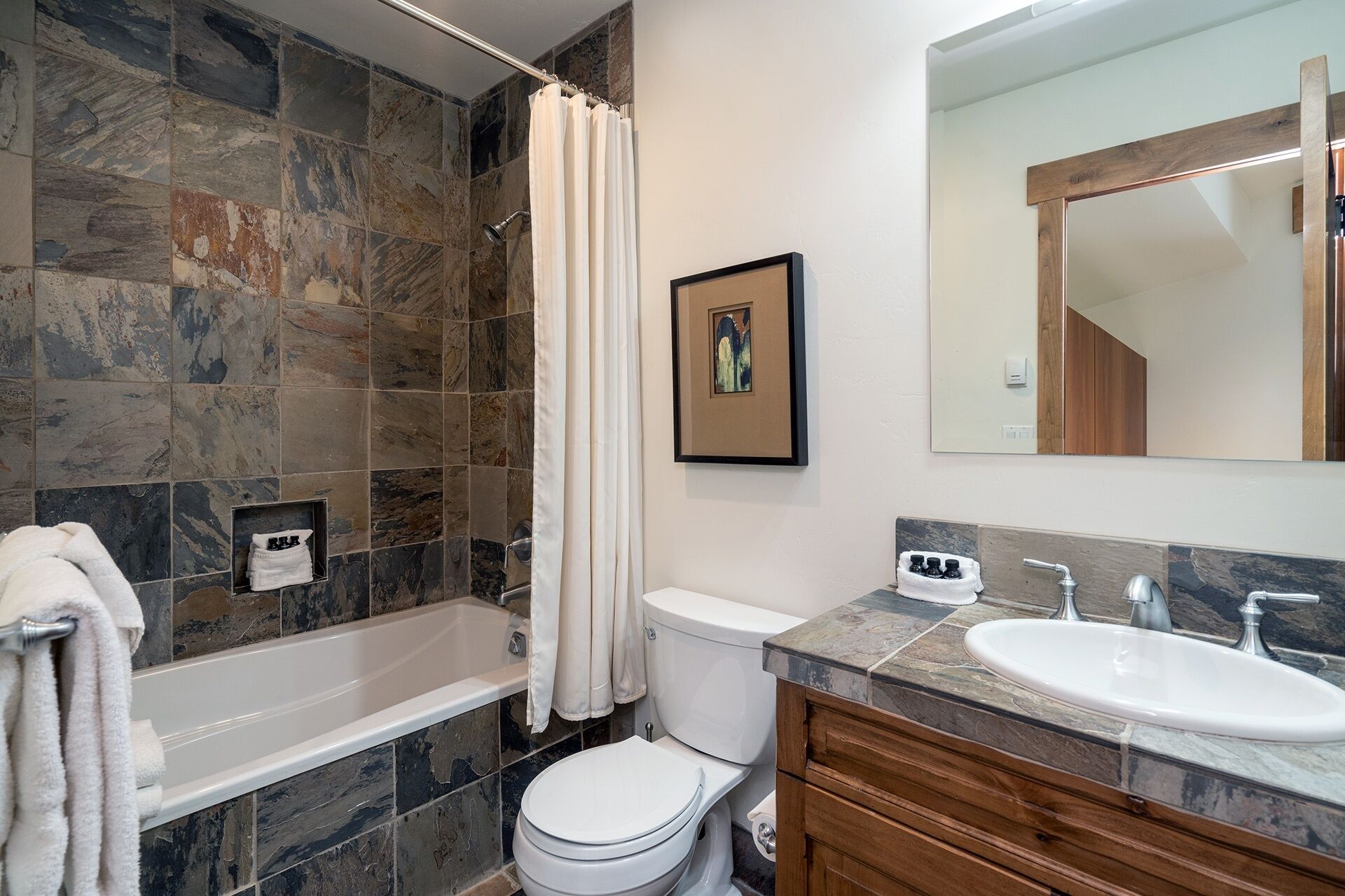 A bathroom with a bathtub, toilet, and sink, featuring stone tiles and a wooden vanity.