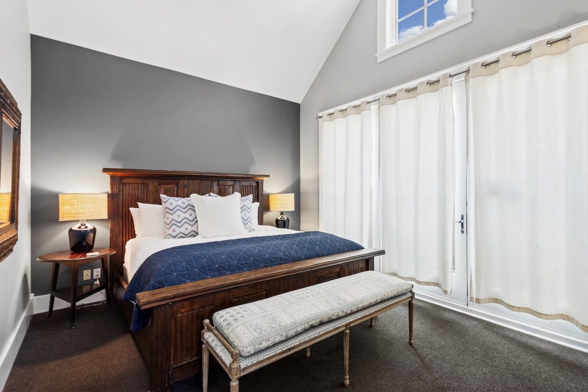 A bedroom with a wooden bed, two bedside lamps, a bench, a large window with curtains, and a mirror on the wall.