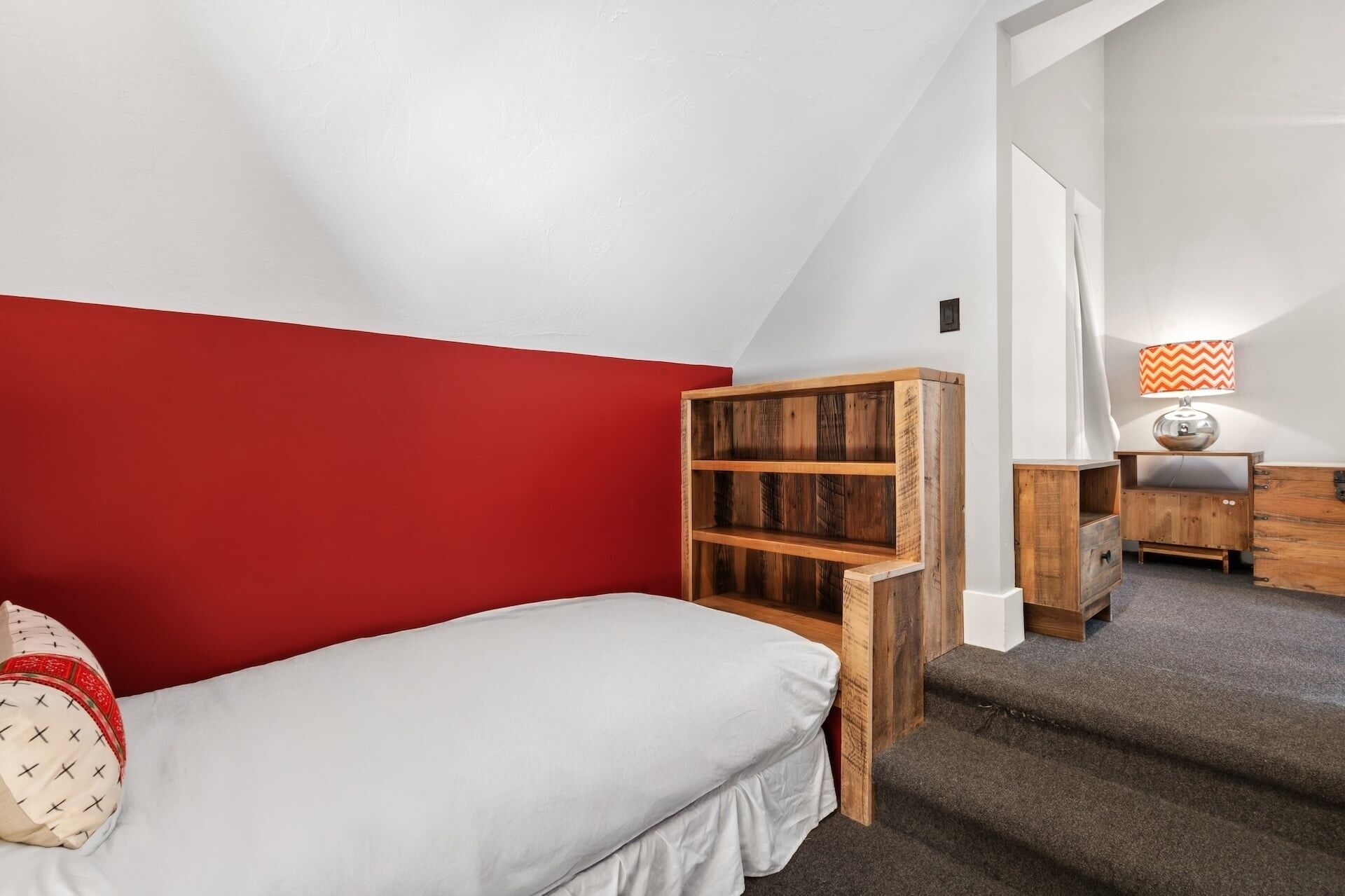A bedroom with a red accent wall, a bed, a wooden bookshelf, a desk, and a lamp.
