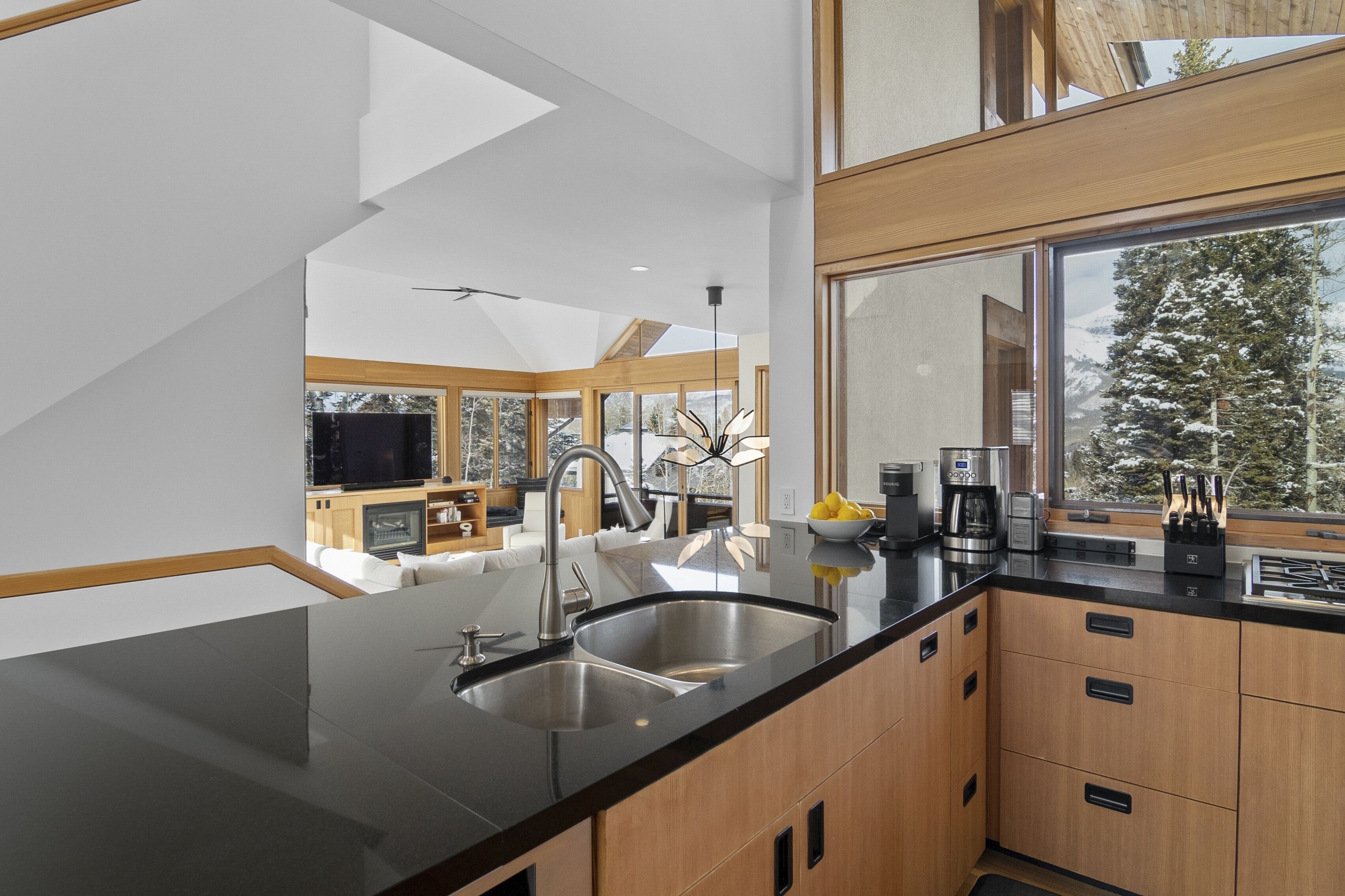 Een moderne keuken met een zwart aanrecht, houten kastjes en een groot raam met uitzicht op besneeuwde bomen.