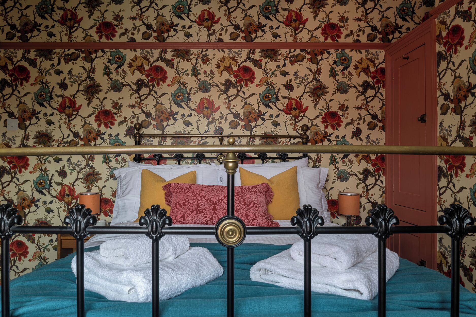 Een bed met bloemenbehang, een metalen hoofdbord en twee witte handdoeken.