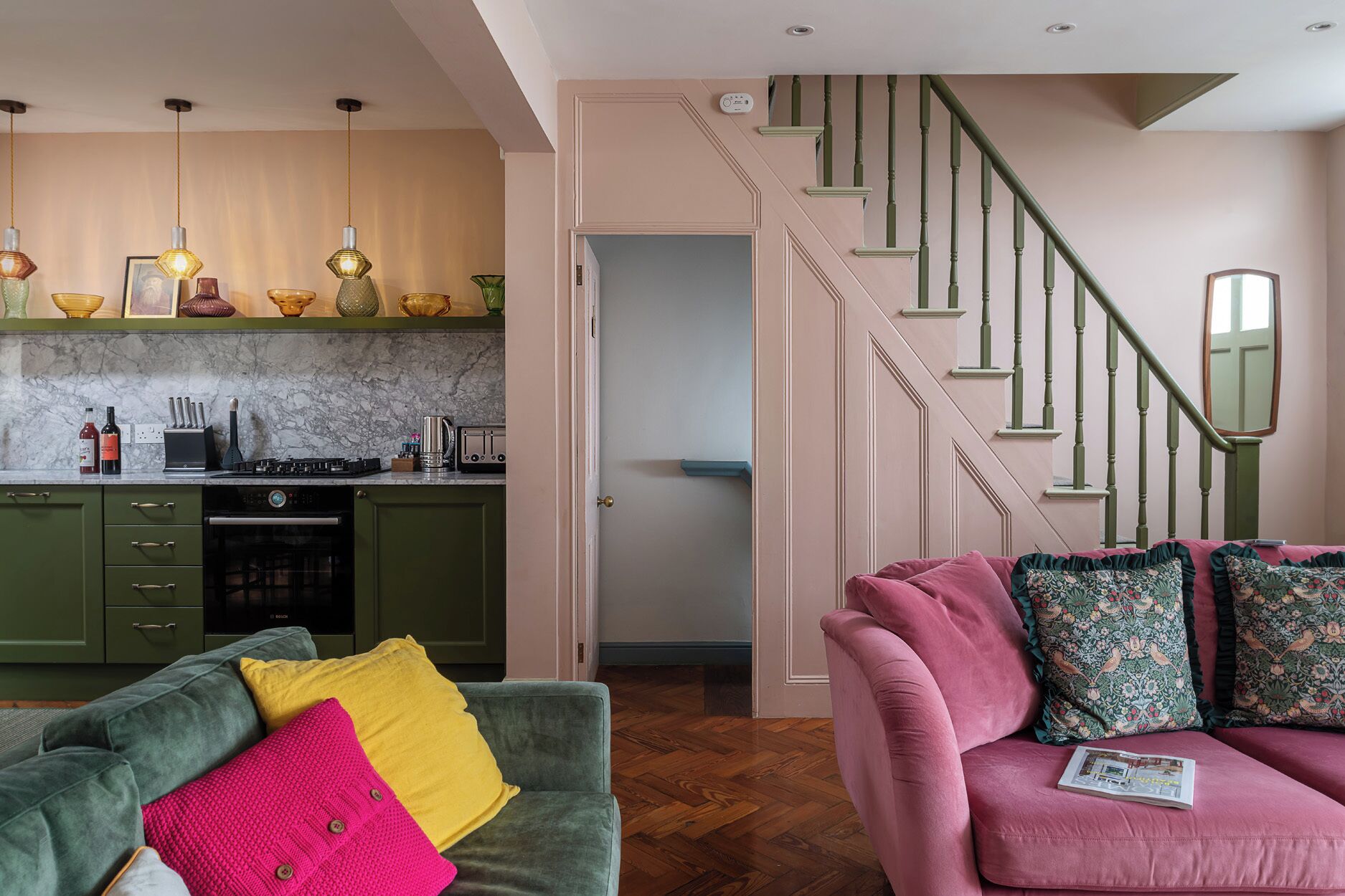 A modern kitchen with pendant lighting, a dining area with a pink sofa and patterned cushions, and a staircase with green railings.