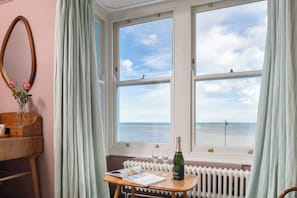 Una habitación con un amplio ventanal con vistas al mar, una mesa de madera con una botella de champán y un espejo en la pared.