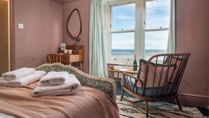 A bedroom with a bed, a chair, a desk, a mirror, and a window with a view of the sea. - Astondene, East Cliff (Reydon)