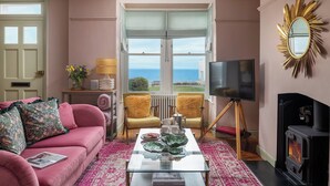 A living room with a sofa, chairs, a coffee table, a fireplace, a television, and a view of the sea. - Astondene, East Cliff (Reydon)