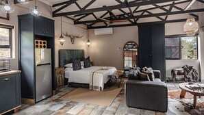 A modern interior with a bed, sofa, and dining area, featuring exposed beams, a large window, and a kitchen with a refrigerator. - Grace of Africa (Nkomazi)