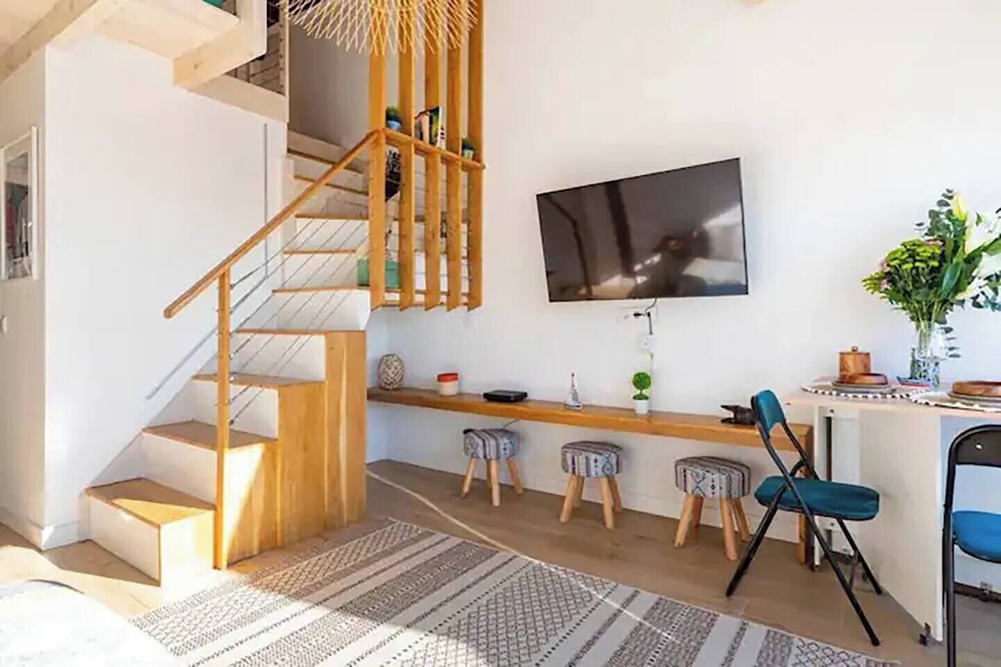 Un espacio habitable moderno con una escalera de madera, un televisor de pantalla plana montado en la pared, una zona de bar con taburetes y una mesa de comedor.