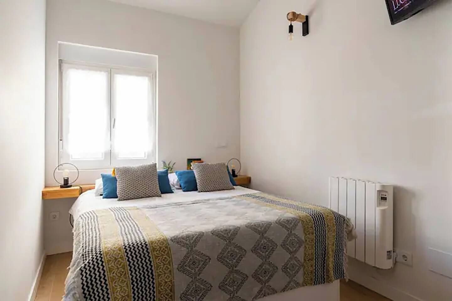 Un dormitorio con cama, ventana, radiador y lámpara fijada a la pared.