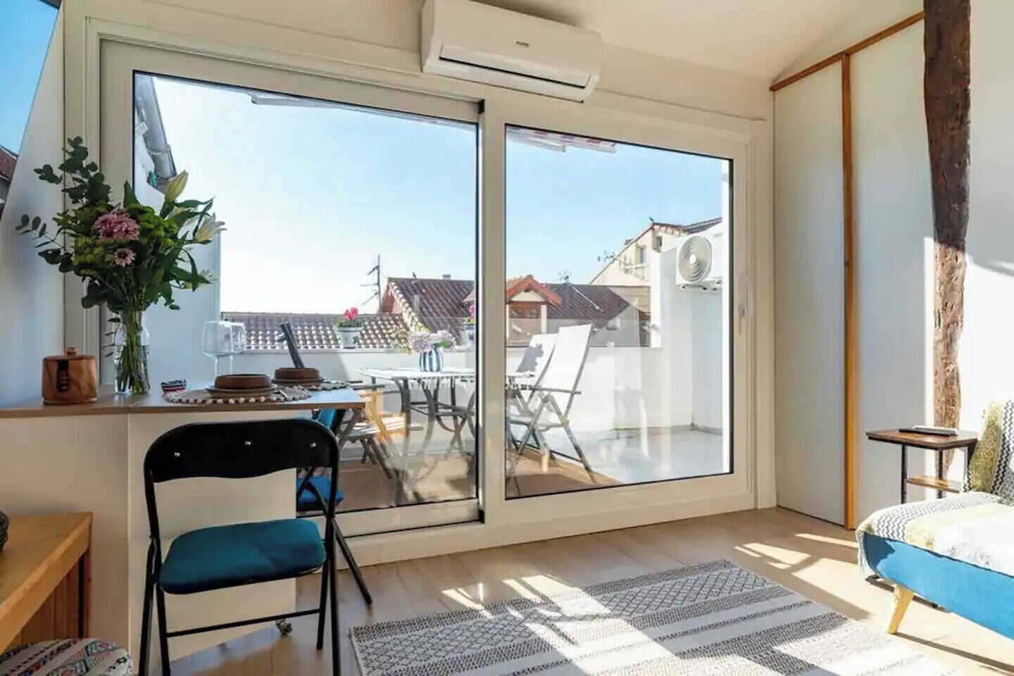 Ein Zimmer mit einer großen Schiebetür, die zu einem Außenbereich führt, einem Esstisch mit Stühlen, einem blauen Sofa und einem kleinen Tisch mit einer Blumenvase.