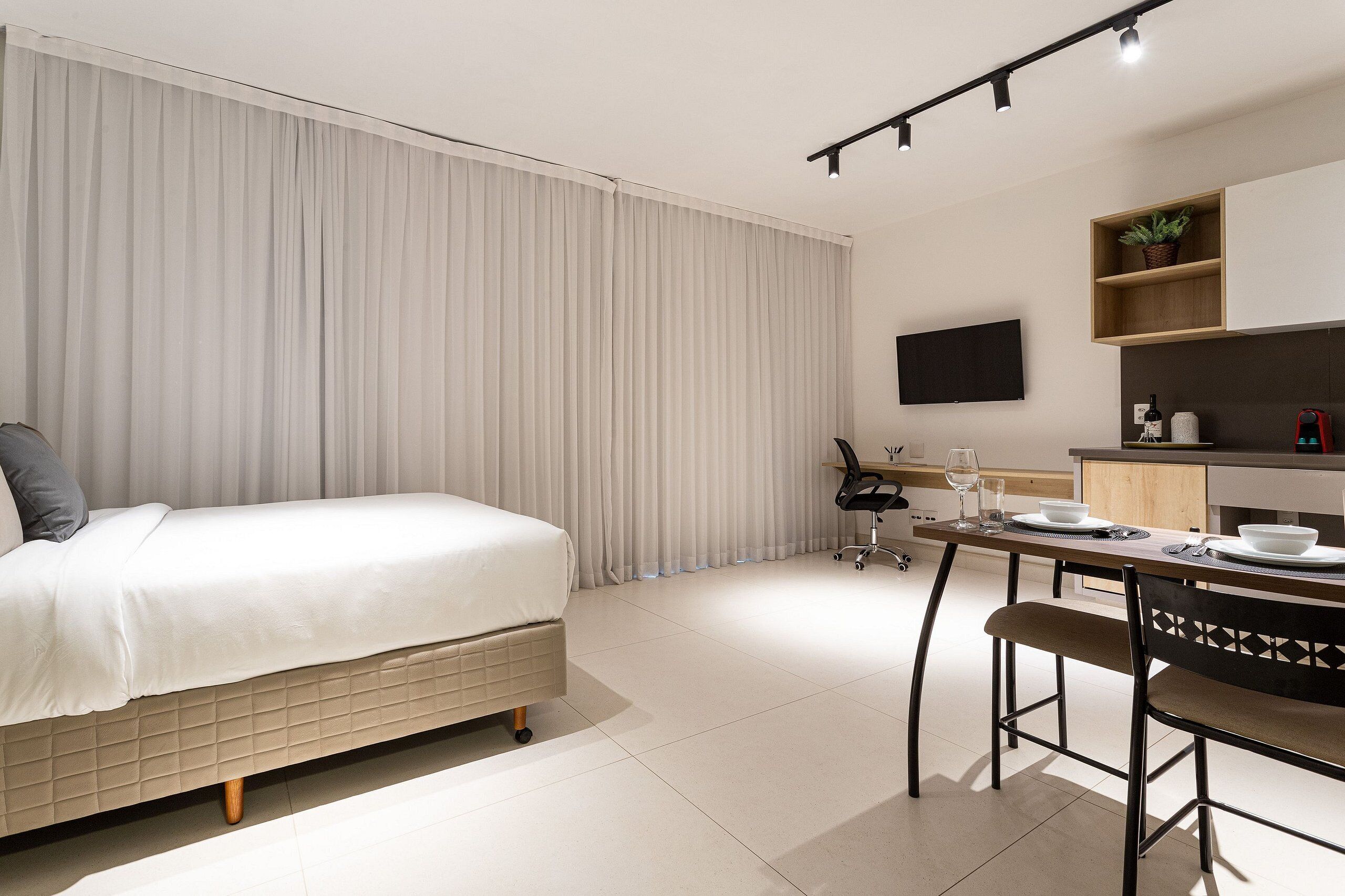 A modern hotel room with a bed, a dining table with chairs, a TV mounted on the wall, and a small kitchen area.
