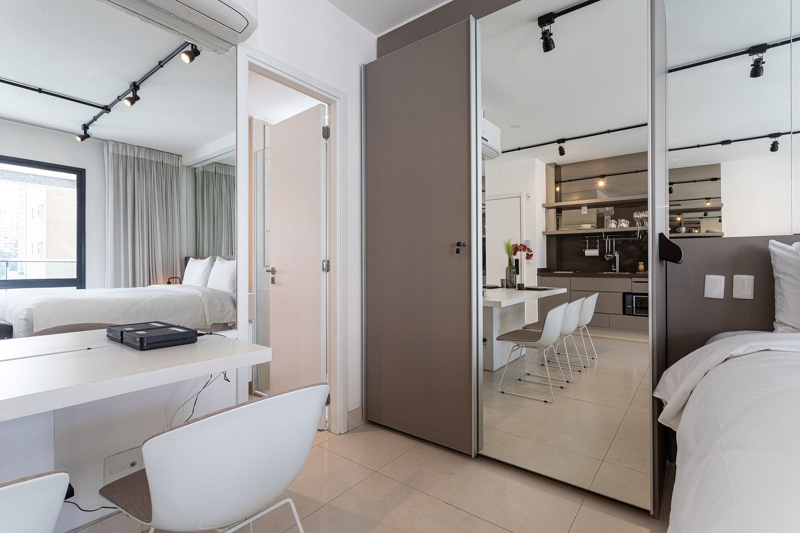 A modern hotel room with a bed, a desk, and a chair. There is a kitchen area visible through an open door.
