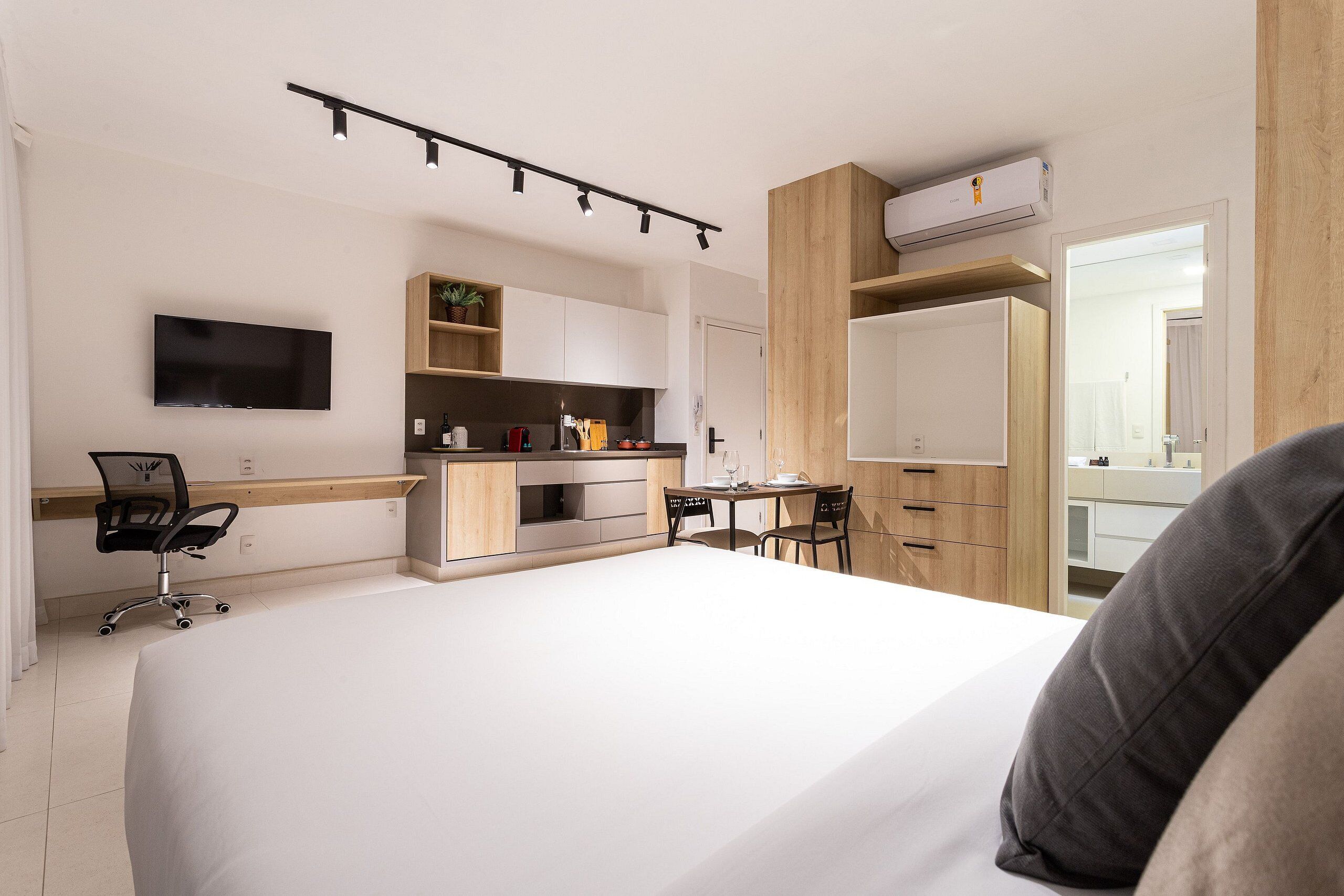 A modern hotel room with a large bed, a desk with a chair, a TV, and a kitchenette.