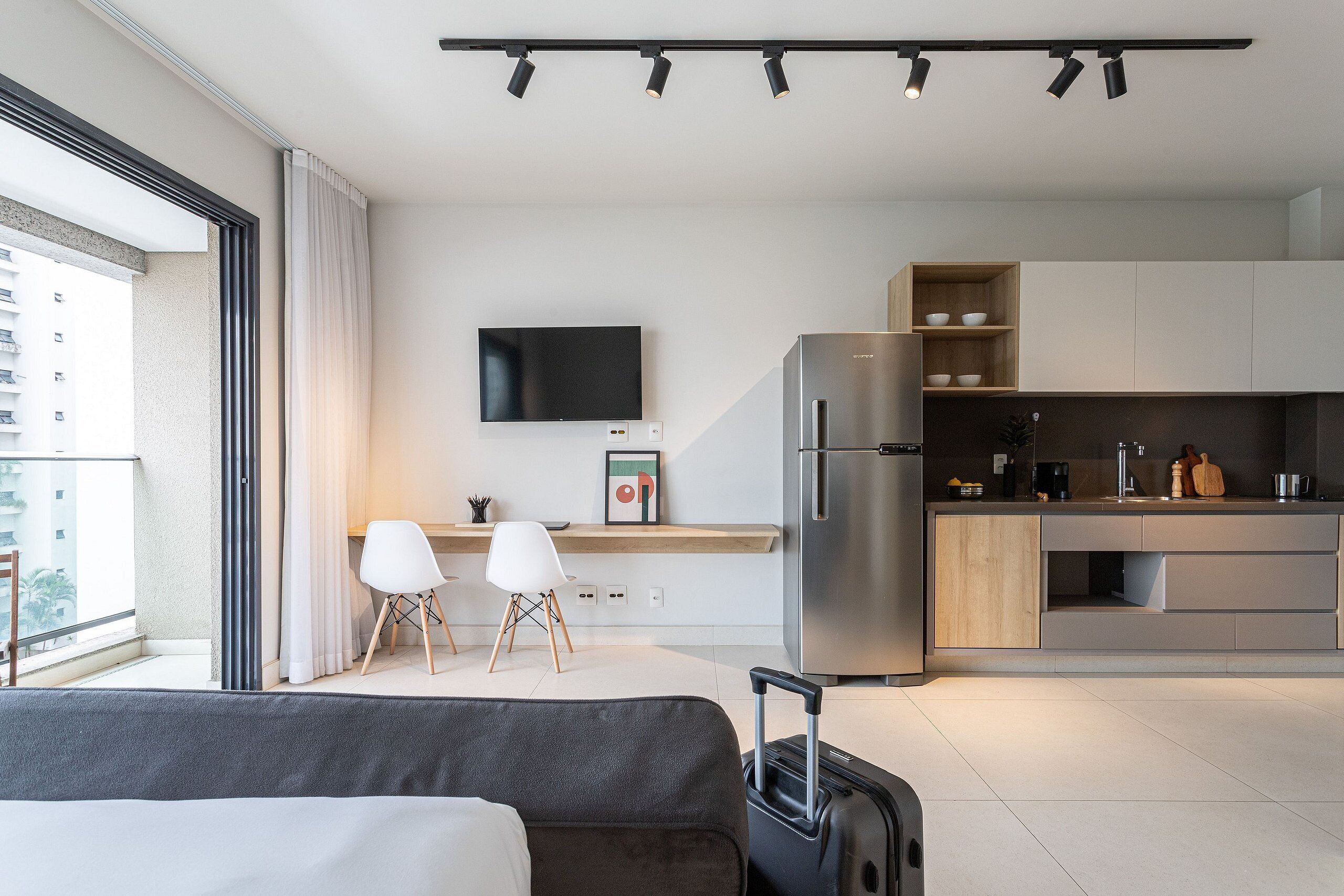 A modern hotel room with a bed, a suitcase, a kitchenette, a flat-screen TV, and a balcony with curtains.