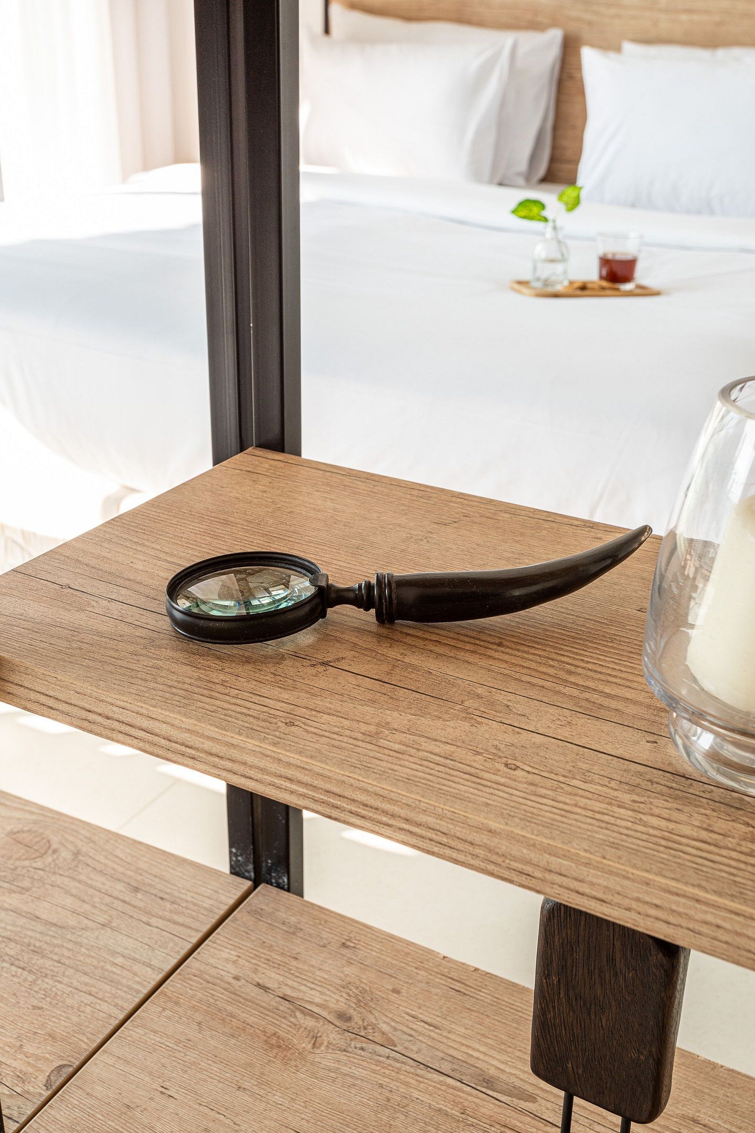 Una mesa de madera con una lupa y un cuenco de vidrio en un balcón, con vistas a una cama con sábanas blancas y una pequeña planta.