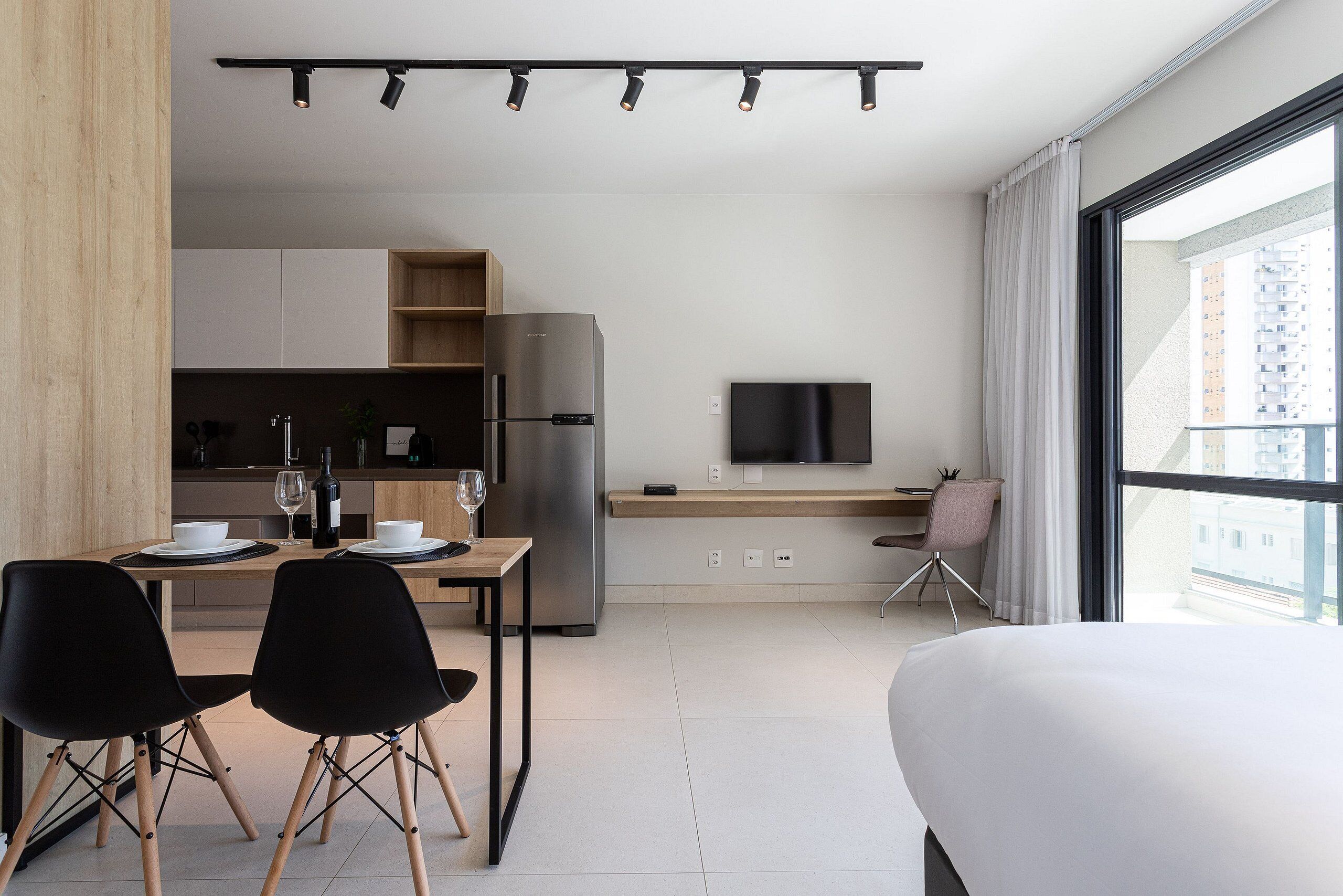 A modern hotel room with a large bed, a dining area with a table and chairs, a kitchen with a refrigerator and a TV, and a view of the city through large windows.